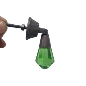 May include: A furniture pull featuring a faceted, translucent green glass drop pendant suspended from a dark, weathered bronze-toned metal mounting bracket. The hardware includes a threaded metal bolt and a nut for secure attachment to a surface.