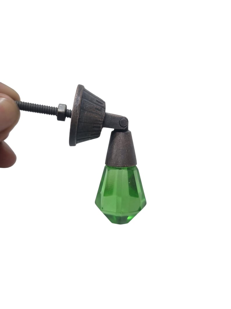 May include: A furniture pull featuring a faceted, translucent green glass drop pendant suspended from a dark, weathered bronze-toned metal mounting bracket. The hardware includes a threaded metal bolt and a nut for secure attachment to a surface.