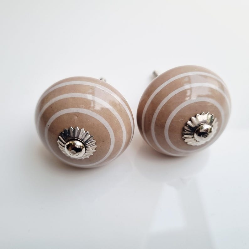 May include: Two decorative cabinet knobs. Each knob is a light brown color with white horizontal stripes. The knobs have a silver-colored, flower-shaped base. These knobs are perfect for adding a touch of style to any cabinet or drawer.