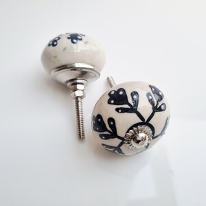 May include: Two ceramic cabinet knobs with a cream-colored base and dark blue floral designs. The knobs have a rounded shape and are mounted on silver-colored metal hardware. The knobs are ideal for adding a decorative touch to furniture.