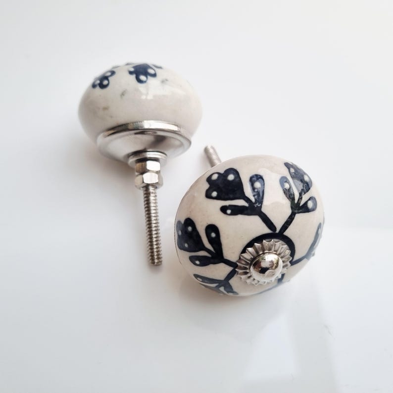 May include: Two ceramic cabinet knobs with a cream-colored base and dark blue floral designs. The knobs have a rounded shape and are mounted on silver-colored metal hardware. The knobs are ideal for adding a decorative touch to furniture.