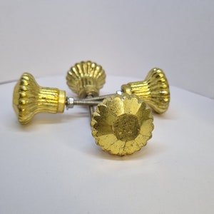 May include: Three gold-toned cabinet knobs with a decorative, scalloped design. The knobs have a shiny finish and are attached to metal screws.