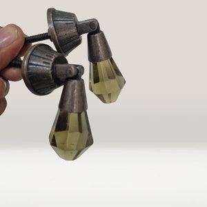 May include: Two drawer pulls featuring a dark bronze-toned metal base with a ribbed, circular mounting plate and a threaded screw. Each pull has a hinged, faceted, translucent olive-green glass drop pendant suspended from the metal hardware.