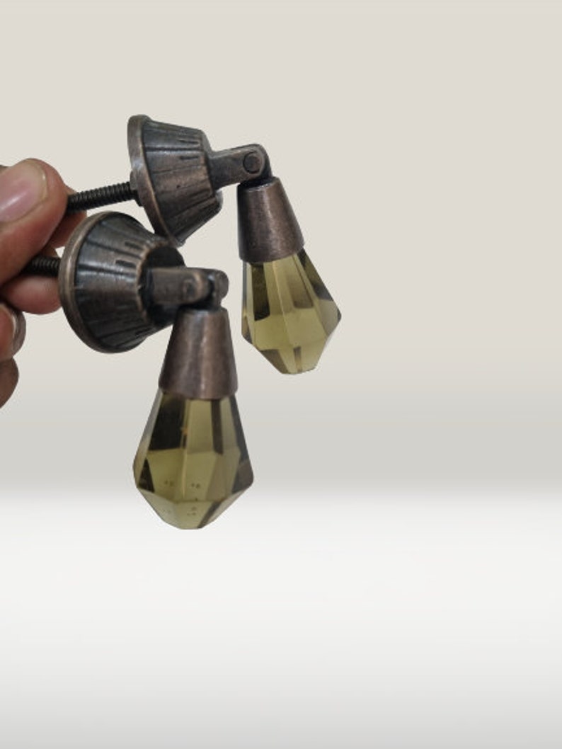 May include: Two drawer pulls featuring a dark bronze-toned metal base with a ribbed, circular mounting plate and a threaded screw. Each pull has a hinged, faceted, translucent olive-green glass drop pendant suspended from the metal hardware.