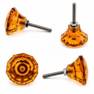 Può includere: Quattro pomelli per armadietti in vetro sfaccettato color ambra con una forma geometrica a più lati. Ogni pomello presenta un bullone in metallo color argento che sporge dal retro, fissato con un dado metallico abbinato per il montaggio.