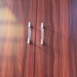 Op de afbeelding: Twee verticale kastgrepen met transparante, cilindervormige acrylhouders en gepolijste zilverkleurige metalen doppen aan de boven- en onderkant, gemonteerd op een donker roodbruin houtnerfoppervlak met een verticale naad tussen de twee grepen.