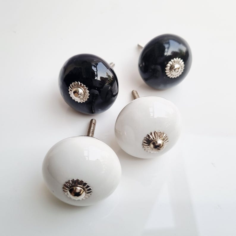 May include: Four decorative cabinet knobs. Two are glossy black, and two are white. Each knob has a silver-colored, flower-shaped base. The knobs are oval-shaped and have a threaded screw for installation.