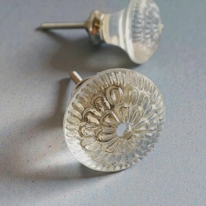 Peut inclure: Deux boutons de placard ronds en verre transparent avec un motif floral texturé en forme de pétales rayonnant à partir d'un trou central. Chaque bouton possède une base en métal argenté et une tige cylindrique courte pour la fixation.