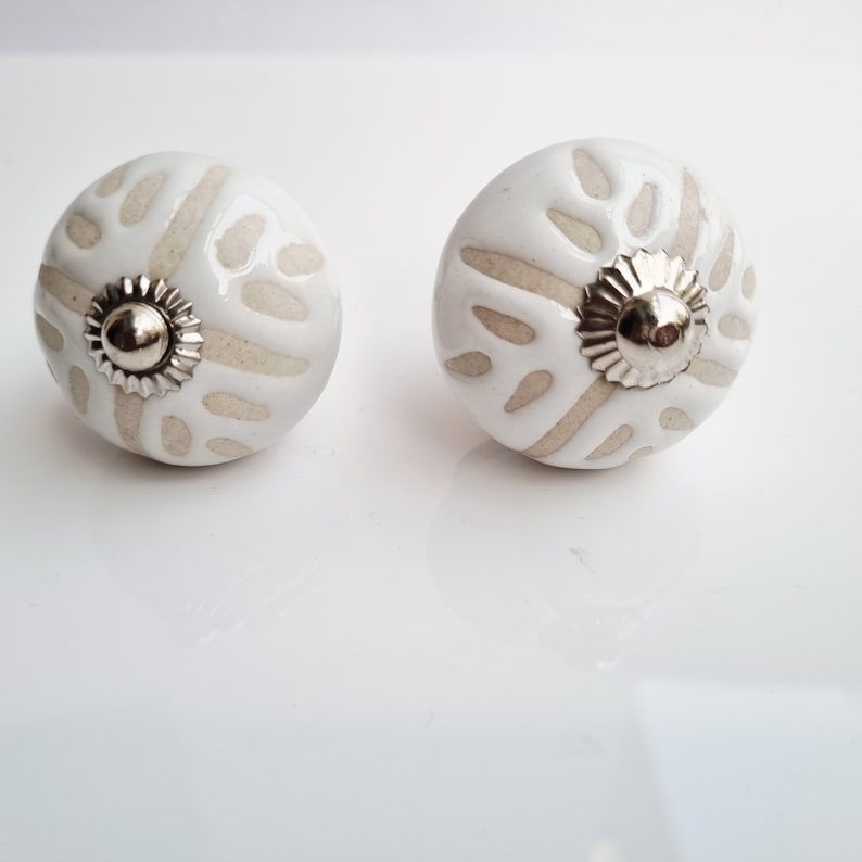 May include: Two white ceramic cabinet knobs with a beige pattern and silver-colored metal accents. The knobs have a round shape and are designed for furniture. The beige pattern is a series of irregular shapes.