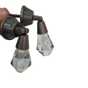 May include: Two drawer pulls featuring a dark bronze-toned metal base with a fluted, conical mounting plate and a hinged, faceted clear glass drop pendant. The hardware includes a threaded metal screw extending from the back of the mounting plate.
