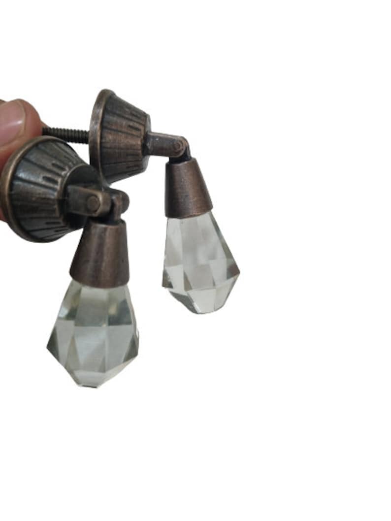 May include: Two drawer pulls featuring a dark bronze-toned metal base with a fluted, conical mounting plate and a hinged, faceted clear glass drop pendant. The hardware includes a threaded metal screw extending from the back of the mounting plate.