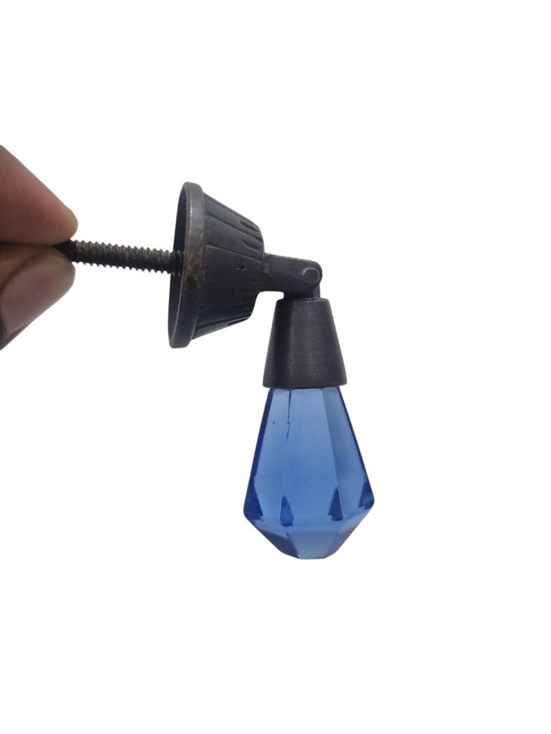 May include: A drop-style furniture pull featuring a faceted, translucent blue glass pendant suspended from a dark, weathered metal mounting bracket. The hardware includes a threaded metal screw stem for attachment to a drawer or cabinet door.