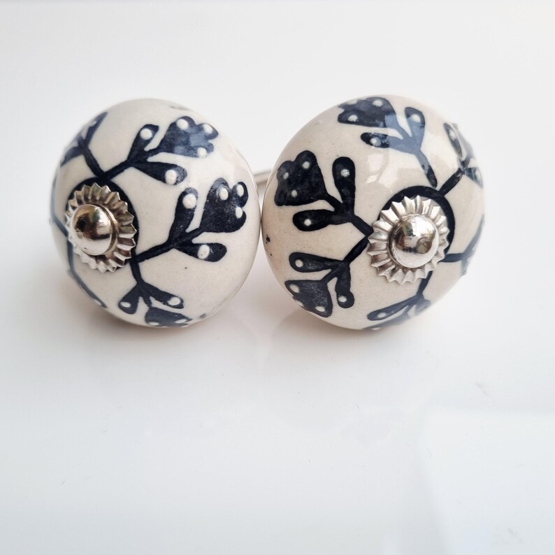 May include: Two round, off-white ceramic cabinet knobs with a dark blue floral design. Each knob has a silver-colored metal base and a matching screw. The knobs are decorative and suitable for furniture.
