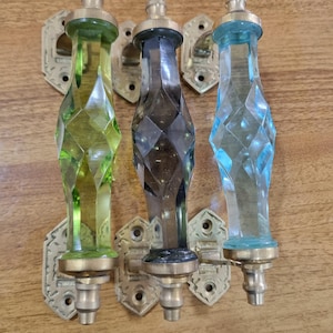 May include: Three decorative glass door handles in green, gray, and blue. Each handle has a faceted design and is mounted on a brass-colored metal plate. The handles are displayed on a wooden surface.