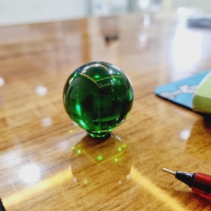 以下が含まれることがあります： 幾何学模様の透明な緑色の球体が正方形の台座に置かれています。球体は光を反射し、赤いペンが前景にあります。オブジェクトは木製の表面にあります。