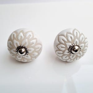 May include: Two white ceramic cabinet knobs with a floral design. The knobs have a silver-colored metal base and are set against a white background. The floral pattern is a lighter shade of white.