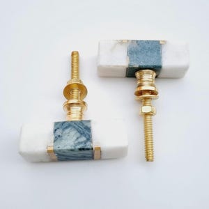 Op de afbeelding: Twee rechthoekige kastknoppen. Elke knop heeft een witte marmeren behuizing met een blauw-grijze gemarmerde middensectie en goudkleurige hardware. De knoppen zijn ontworpen voor eenvoudige installatie.