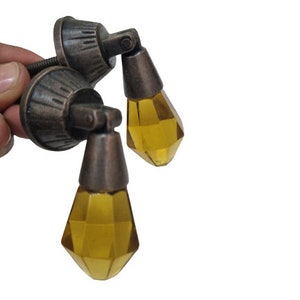 May include: Two drop-style cabinet pulls featuring faceted, amber-colored glass pendants suspended from antique bronze-finished metal mounts. Each mount has a circular base with a ribbed texture and a hinged mechanism that allows the glass to hang.