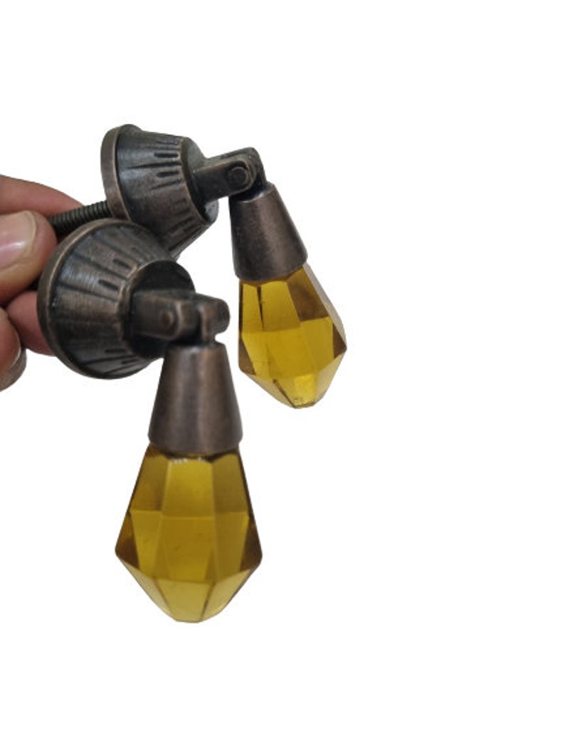 May include: Two drop-style cabinet pulls featuring faceted, amber-colored glass pendants suspended from antique bronze-finished metal mounts. Each mount has a circular base with a ribbed texture and a hinged mechanism that allows the glass to hang.