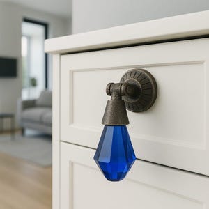 May include: A decorative cabinet pull featuring a faceted, teardrop-shaped blue glass pendant suspended from a circular, antique bronze-toned metal mounting plate with a radial grooved pattern, attached to a white wooden drawer front.