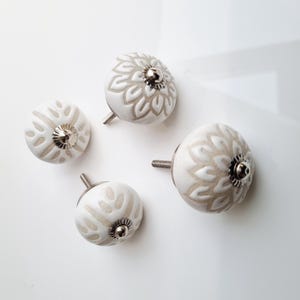 May include: Four white ceramic cabinet knobs with a floral design in a light beige color. Each knob has a silver-colored metal base and screw. The knobs are in various sizes and are arranged on a white surface.