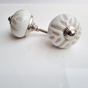 May include: Two white ceramic cabinet knobs with a floral design in a neutral color. The knobs have a silver metal base and screw. The knobs are a decorative element for furniture.