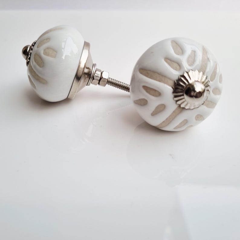 May include: Two white ceramic cabinet knobs with a floral design in a neutral color. The knobs have a silver metal base and screw. The knobs are a decorative element for furniture.