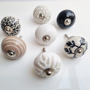 May include: A collection of eight decorative ceramic cabinet knobs. The knobs feature various designs, including floral patterns, stripes, and solid colors like white and black. Each knob has a silver-toned metal base and stem for mounting.