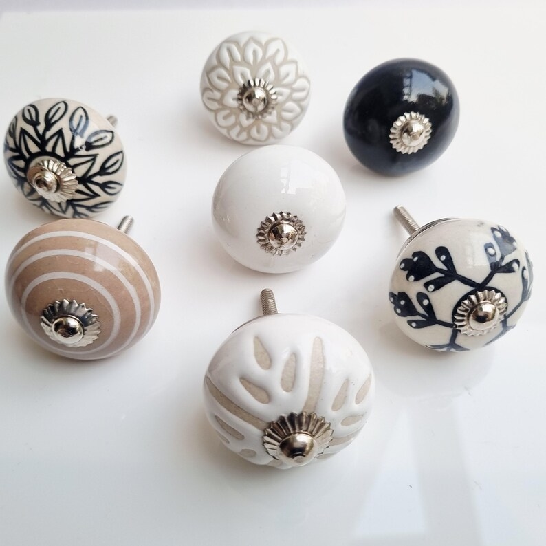 May include: A collection of eight decorative ceramic cabinet knobs. The knobs feature various designs, including floral patterns, stripes, and solid colors like white and black. Each knob has a silver-toned metal base and stem for mounting.