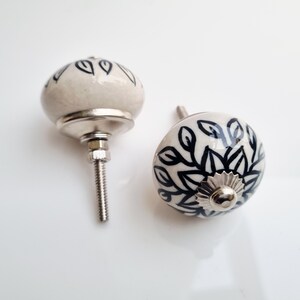 May include: Two ceramic cabinet knobs with a white base and black leaf design. The knobs have a rounded shape and are attached to a silver metal base with a threaded screw for installation. The knobs are decorative and suitable for furniture.
