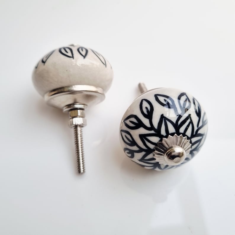 May include: Two ceramic cabinet knobs with a white base and black leaf design. The knobs have a rounded shape and are attached to a silver metal base with a threaded screw for installation. The knobs are decorative and suitable for furniture.