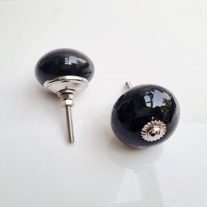 May include: Two black ceramic cabinet knobs with a glossy finish. Each knob has a silver-colored base and a threaded screw for installation. The knobs are oval-shaped and reflect light.