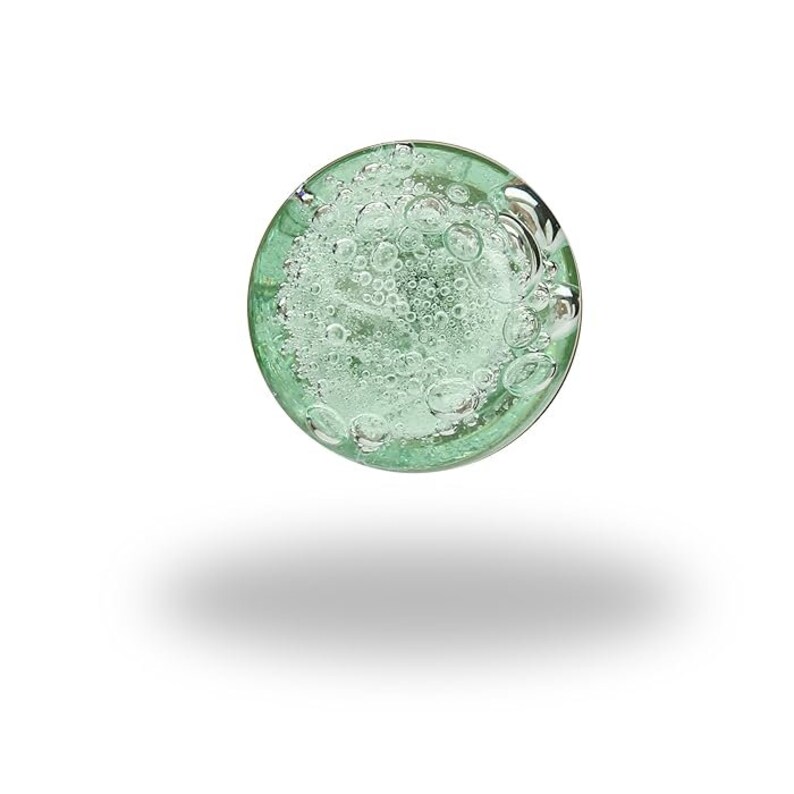 May include: A circular glass paperweight featuring a translucent light green tint. The interior of the glass is filled with numerous small, suspended air bubbles of varying sizes, creating a textured, effervescent appearance throughout the sphere.