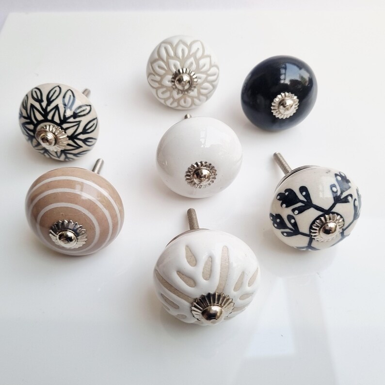 May include: A collection of seven ceramic cabinet knobs in various designs and colors. The knobs feature floral, striped, and solid patterns in white, black, gray, and tan. Each knob has a silver-toned metal base and screw.