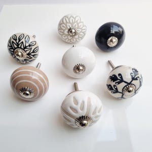 May include: A collection of seven ceramic cabinet knobs in various designs and colors. The knobs feature floral, striped, and solid patterns in white, black, beige, and gray. Each knob has a silver-toned metal base and screw.