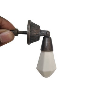 May include: A cabinet knob featuring a white, faceted, diamond-shaped ceramic pull suspended from a dark, antique-bronze metal base. The hardware includes a threaded metal bolt and a small nut, designed for attachment to furniture or cabinetry doors.
