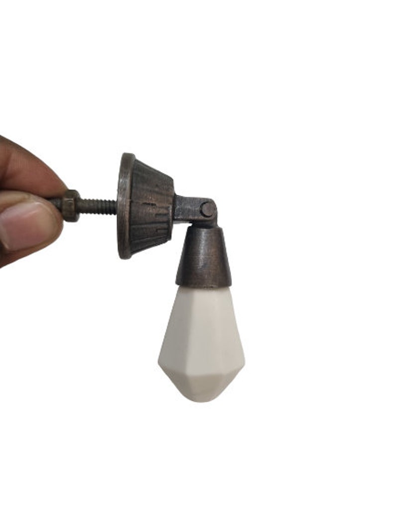 May include: A cabinet knob featuring a white, faceted, diamond-shaped ceramic pull suspended from a dark, antique-bronze metal base. The hardware includes a threaded metal bolt and a small nut, designed for attachment to furniture or cabinetry doors.