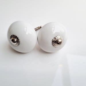 May include: Two white ceramic cabinet knobs with a glossy finish. Each knob features a silver-colored, flower-shaped base. The knobs are oval-shaped and have a simple, elegant design, suitable for various furniture styles.