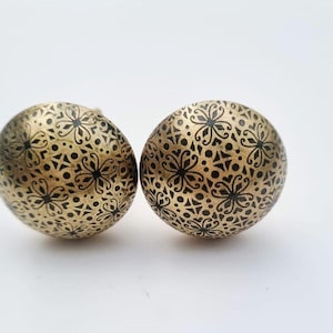 Op de afbeelding: Twee decoratieve, goudkleurige kastknoppen. Elke knop is rond met een gedetailleerd zwart bloemenpatroon. De knoppen zijn gemaakt van metaal en staan tegen een witte achtergrond. Deze knoppen zijn ideaal om een vleugje elegantie aan meubels toe te voegen.