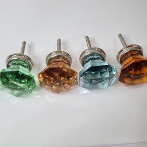 May include: Four glass cabinet knobs with a faceted diamond shape. The knobs are in shades of green, brown, blue, and amber. Each knob has a silver metal base and a screw.