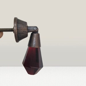 May include: A drop-style cabinet pull featuring a faceted, deep red glass pendant suspended from a dark, antique-finished metal mounting base. The hardware includes a threaded metal screw extending from the back for secure installation.