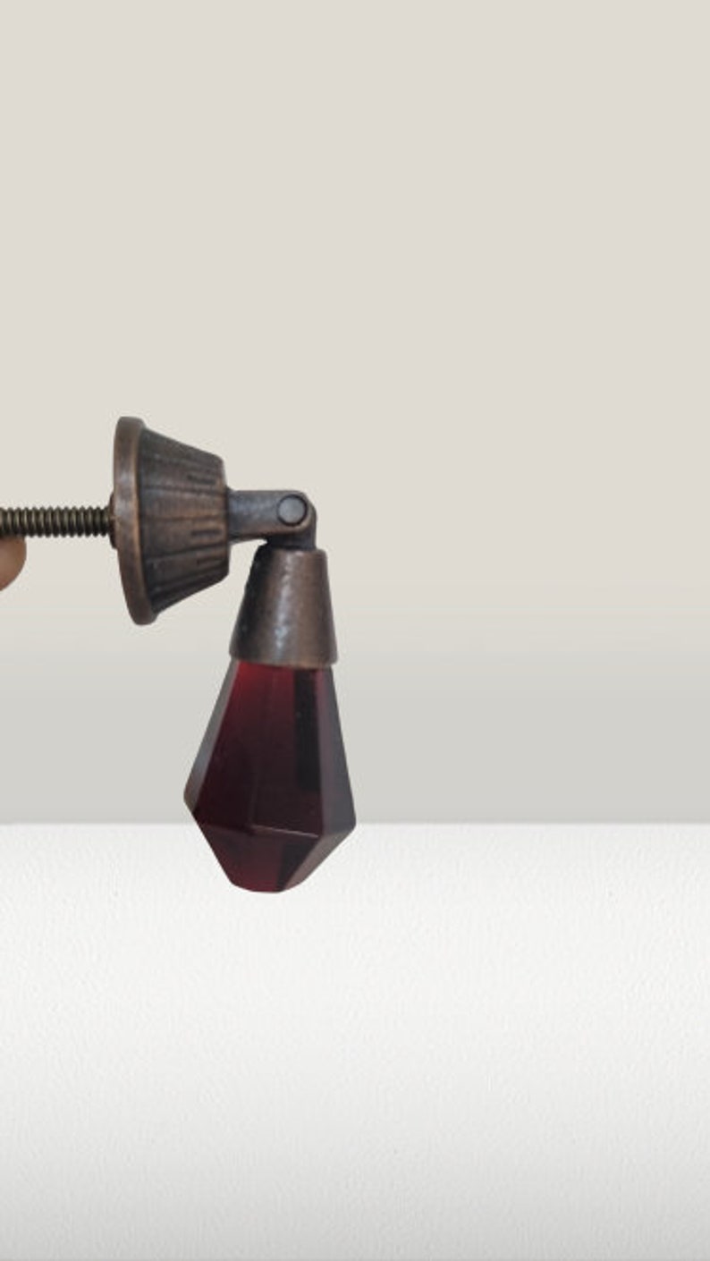 May include: A drop-style cabinet pull featuring a faceted, deep red glass pendant suspended from a dark, antique-finished metal mounting base. The hardware includes a threaded metal screw extending from the back for secure installation.