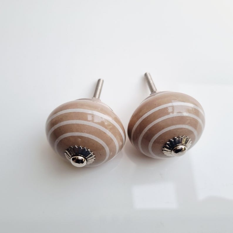 May include: Two decorative cabinet knobs. Each knob is a light brown color with white horizontal stripes. The knobs have a silver-colored base and a threaded post for installation. The knobs are designed for use on drawers or cabinets.