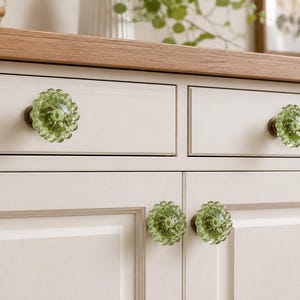 May include: Four round, light green glass cabinet knobs with a textured, scalloped edge design and a dark metal base. These decorative knobs are attached to a light-colored wooden cabinet featuring two upper drawers and two lower cabinet doors.