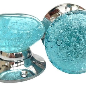 May include: Two decorative glass door knobs. The knobs are a light blue color with a bubble-like texture. The base of the knobs is a shiny silver color. The knobs are designed for use on doors or drawers.