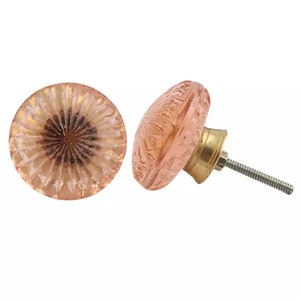 Op de afbeelding: Twee ronde kastknoppen van doorschijnend lichtroze glas met een textuur in een straalsgewijs patroon. Elke knop heeft een donkerbruin accent in het midden en is bevestigd aan een goudkleurige metalen voet met een metalen schroefdraad.