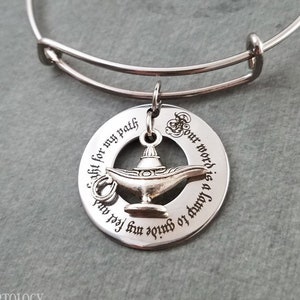 May include: A silver bangle bracelet with a charm featuring a genie lamp and the inscription "Pour word is a lamp to guide my feet and light for my path".