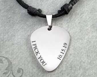 May include: A silver guitar pick pendant necklace with the words "I PICK YOU" and the date "10.15.19" engraved on it. The pendant hangs from a black leather cord.