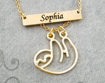 May include: Gold-colored necklace featuring a rectangular bar engraved with the name "Sophia" and a dangling sloth pendant. The necklace has a delicate chain and is set against a gray background.