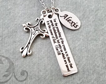 May include: Silver necklace featuring a cross charm, a rectangular charm inscribed with the text "For the preaching of the cross is to them that perish foolishness; but unto us which are saved it is the power of God. I Cor.1:18" and a round charm engraved with the name "Alexis".